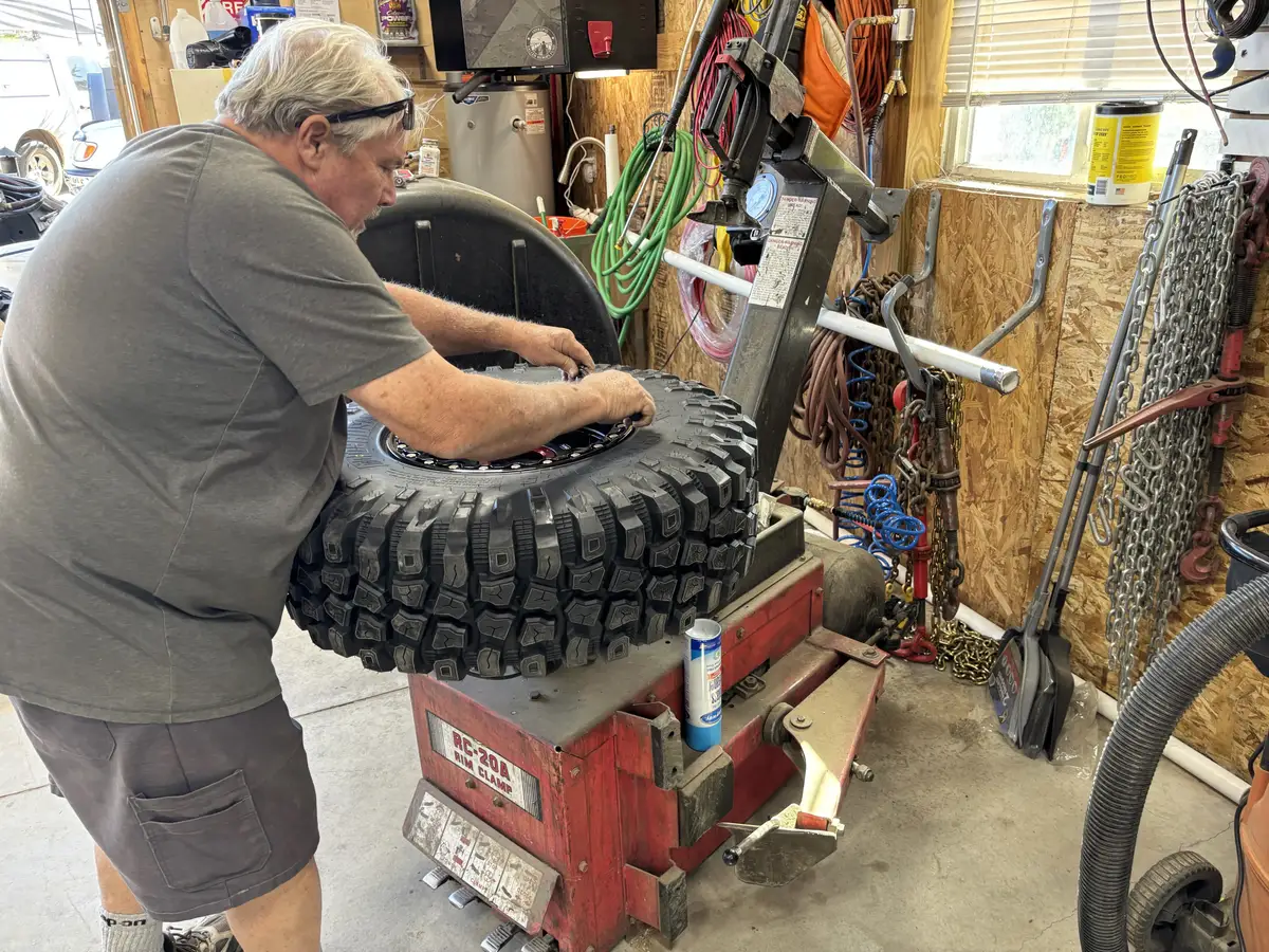 Seating the bead of a large off-road tire at the tire machine, with chains and tools racked on the wall behind